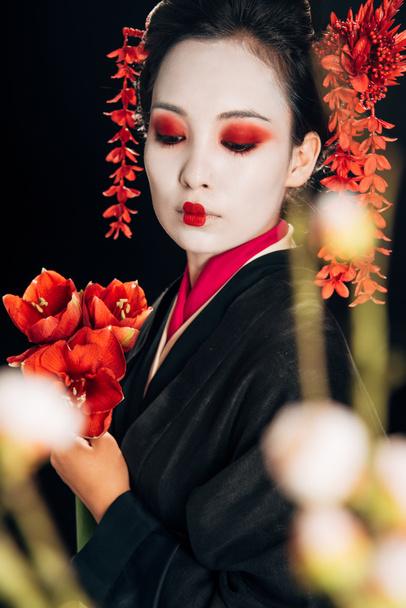 selective focus of beautiful geisha in black kimono with red flowers and sakura branches isolated on black - Fotografie, Obrázek