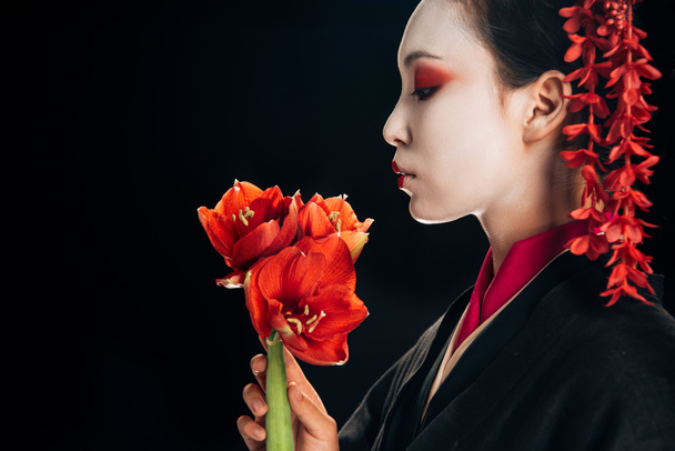side view of beautiful geisha in black kimono with red flowers isolated on black - Photo, Image
