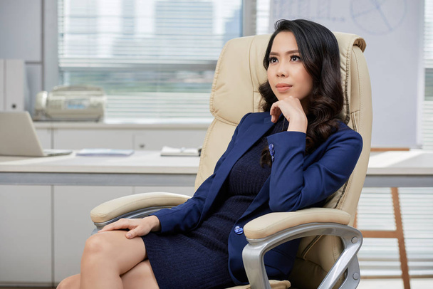 Joven mujer de negocios asiática sentada en silla cómoda en la oficina y pensando en algo
 - Foto, Imagen