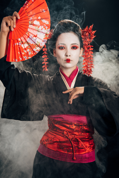 beautiful geisha in black kimono with flowers in hair holding hand fan and gesturing in smoke - Photo, Image