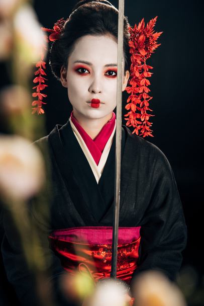selective focus of confident geisha in black kimono holding katana and sakura branches isolated on black - Photo, Image