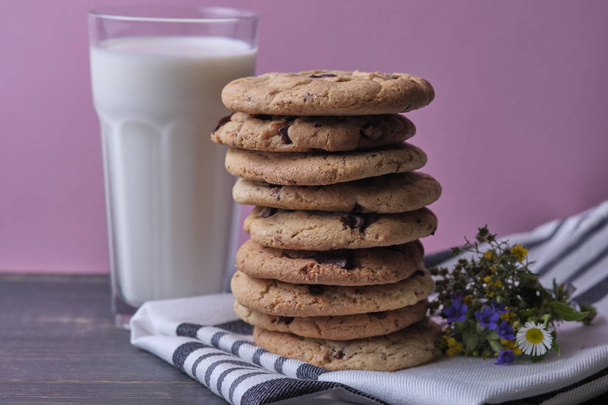 Αμερικάνικα μπισκότα σοκολάτας, ένα ποτήρι γάλα και αγριολούλουδα. Σκούρο ξύλινο τραπέζι και ροζ φόντο. - Φωτογραφία, εικόνα