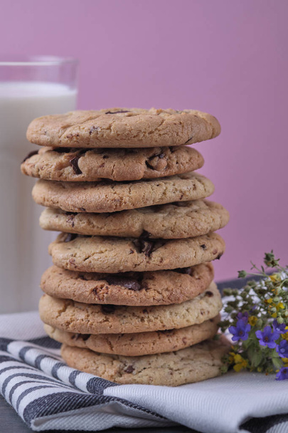 Αμερικάνικα μπισκότα σοκολάτας, ένα ποτήρι γάλα και αγριολούλουδα. Σκούρο ξύλινο τραπέζι και ροζ φόντο. - Φωτογραφία, εικόνα