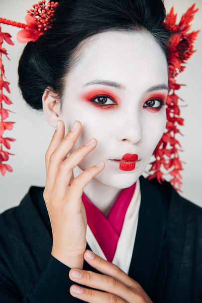 portrait of beautiful geisha in black kimono with red flowers in hair touching face isolated on white - 写真・画像