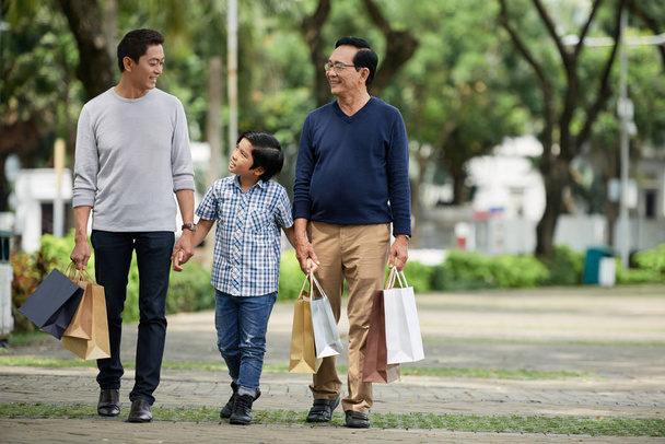 Sweet boy in casual outfit walking along street path with smiling father and grandfather after shopping on sunny day - Photo, Image
