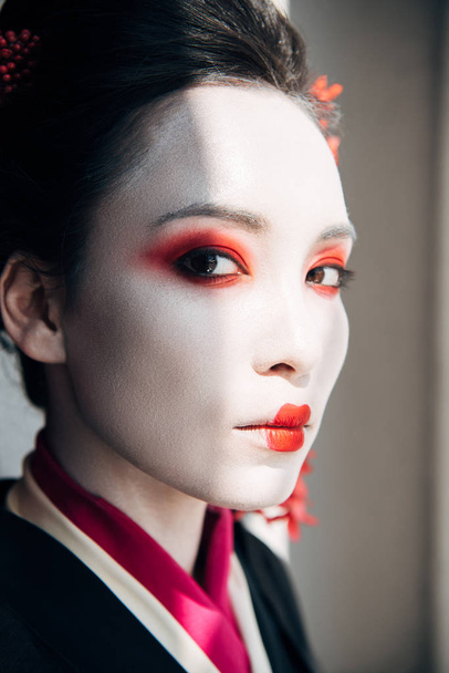 portrait of beautiful geisha with red and white makeup looking at camera in sunlight - Φωτογραφία, εικόνα