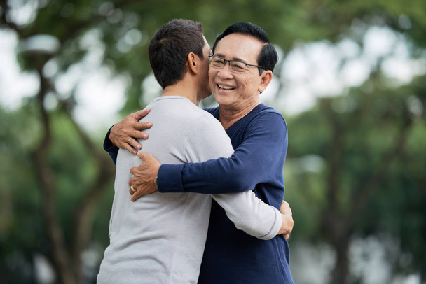 Пожилой азиат улыбается и обнимает сына стоя на размытом фоне парка
 - Фото, изображение