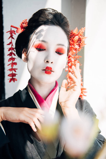 selective focus of sakura branches and beautiful geisha with red and white makeup and closed eyes in sunlight - Photo, Image