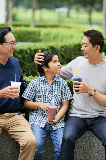 Adorabile asiatico ragazzo holding cup di yummy drink e sorridente mentre seduta a parco vicino padre e nonno
 - Foto, immagini