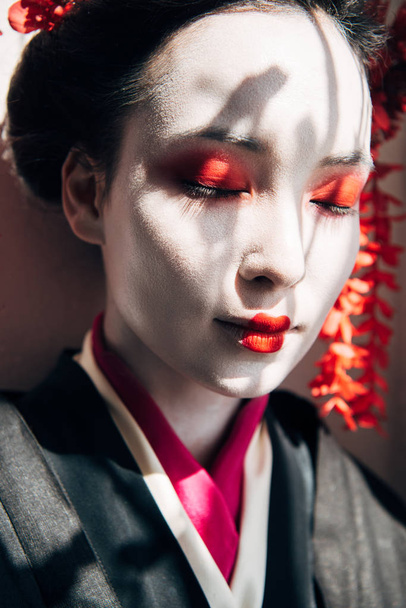 πορτρέτο της όμορφης γκέισα με κόκκινο και λευκό μακιγιάζ και κλειστά μάτια στο ηλιακό φως με σκιές - Φωτογραφία, εικόνα