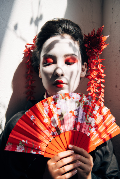 Porträt der wunderschönen Geisha mit rot-weißem Make-up, die einen Fächer im Sonnenlicht hält - Foto, Bild
