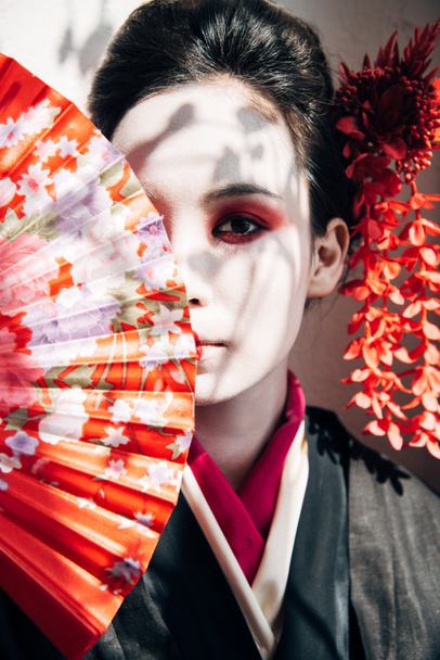 belle geisha avec maquillage rouge et blanc tenant ventilateur de main près du visage dans la lumière du soleil
 - Photo, image