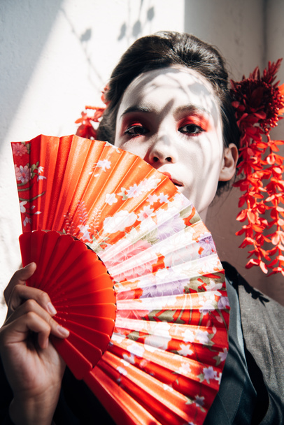 portrait of beautiful geisha with red and white makeup holding hand fan in sunlight - Foto, Imagen