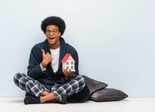 Joven hombre negro sosteniendo un modelo de casa sentado en el suelo sorprendido, se siente exitoso y próspero
 - Foto, imagen