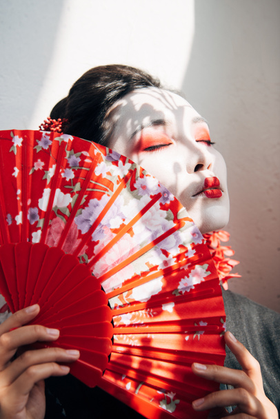 beautiful geisha with red and white makeup and closed eyes holding hand fan in sunlight - Фото, зображення