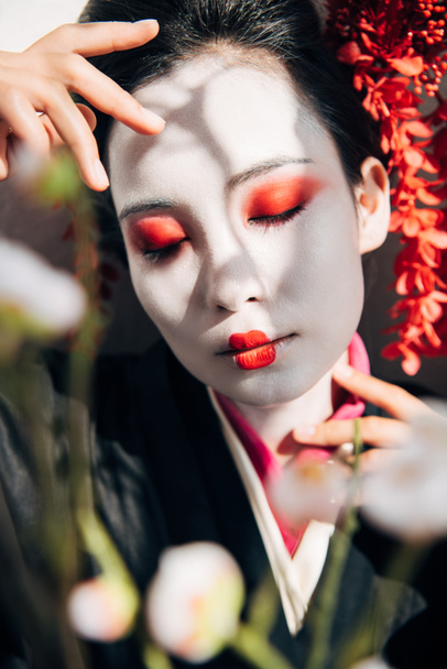 selective focus of tree branches and beautiful geisha with red and white makeup and closed eyes in sunlight - Fotó, kép