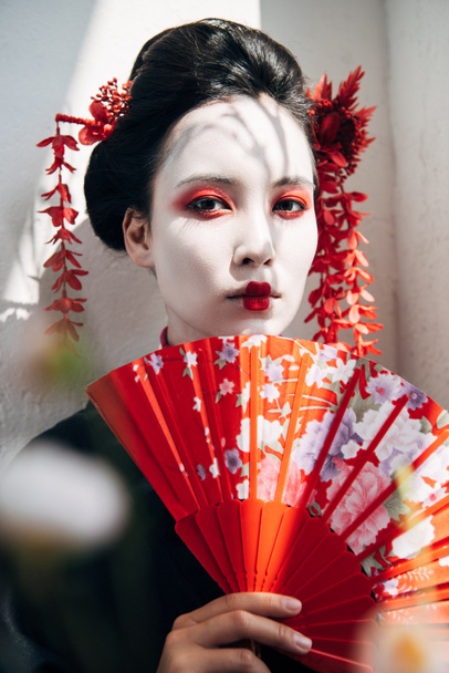 selektiver Fokus von Baumzweigen und wunderschöner Geisha mit rot-weißem Make-up, die einen Handfächer im Sonnenlicht hält - Foto, Bild