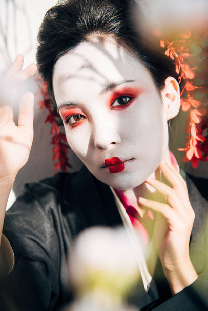 selective focus of tree branches and beautiful geisha with red and white makeup in sunlight - Fotoğraf, Görsel