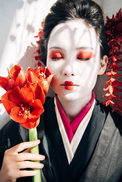 portrait of beautiful geisha holding red flowers in sunlight - Photo, Image