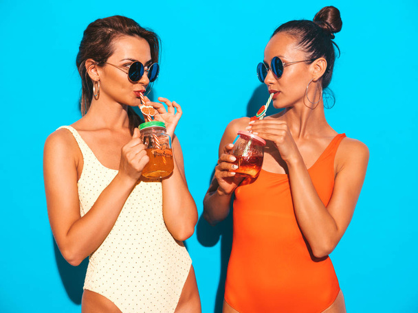 Two beautiful sexy smiling hipster women in summer colorful swimwear bathing suits.Trendy girls in sunglasses.Going crazy.Funny models isolated on blue.Drinking fresh cocktail smoozy drink - 写真・画像