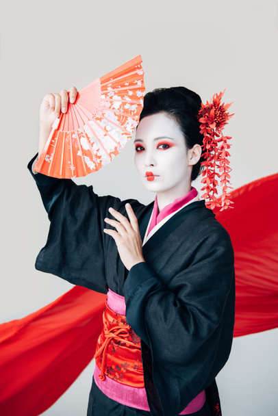hermosa geisha en kimono negro posando con ventilador de mano y tela roja sobre fondo aislado en blanco
 - Foto, Imagen