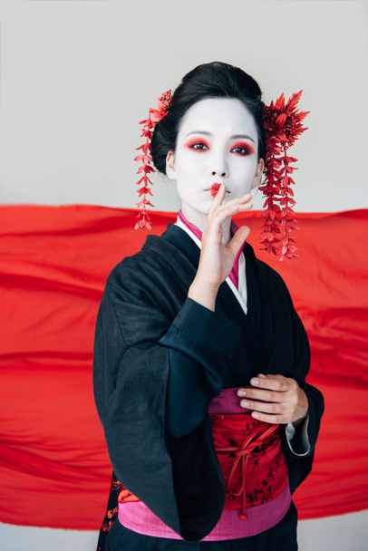 красивая гейша в черном кимоно с красной тканью на заднем плане, показывающая знак "ш", изолированный на белом
 - Фото, изображение