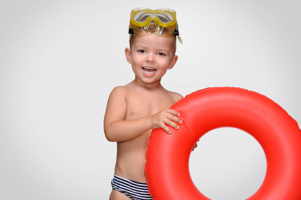 Netter kleiner Junge lacht in gestreifter Badehose mit rotem aufblasbarem Ring und Taucherbrille - Foto, Bild
