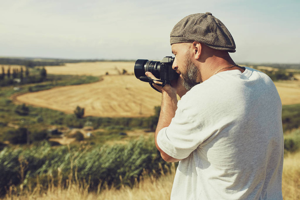 photographer with a camera stands on the background of cereal fields. a man wearing shorts and a t-shirt, a cap on his head - Photo, Image