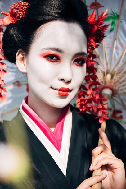 selective focus of beautiful smiling geisha with umbrella in sunlight - Photo, Image