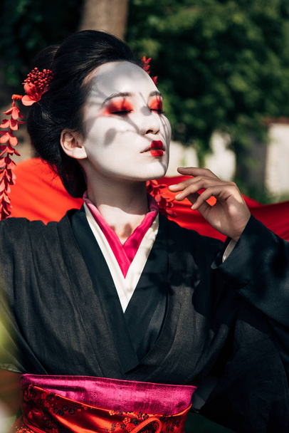 selectieve focus van boomtakken en mooie Geisha met gesloten ogen en rode doek op achtergrond in zonlicht - Foto, afbeelding