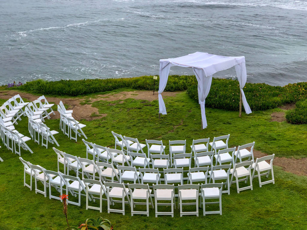Свадебная церемония установки с белыми стульями и аркой в саду перед океаном, свадебная концепция, Ла-Хойя, Калифорния, США
 - Фото, изображение