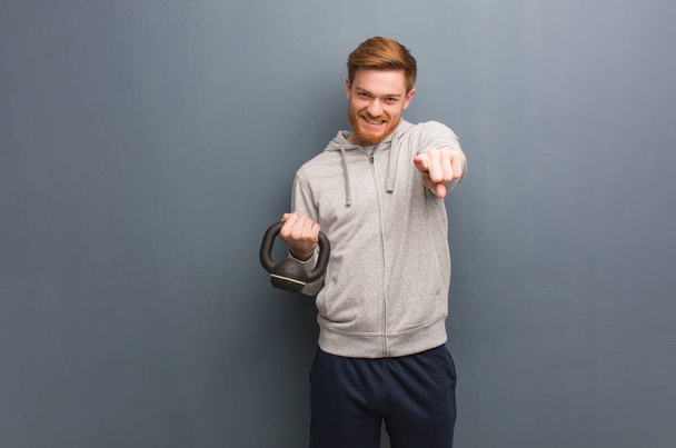 Jeune homme de fitness rousse joyeux et souriant pointant vers l'avant. Tenant un haltère
. - Photo, image