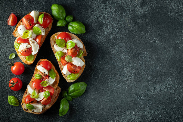 Bruschetta with tomatoes, mozzarella cheese and basil on a dark background. Traditional italian appetizer or snack, antipasto. Top view with copy space. Flat lay - Photo, Image