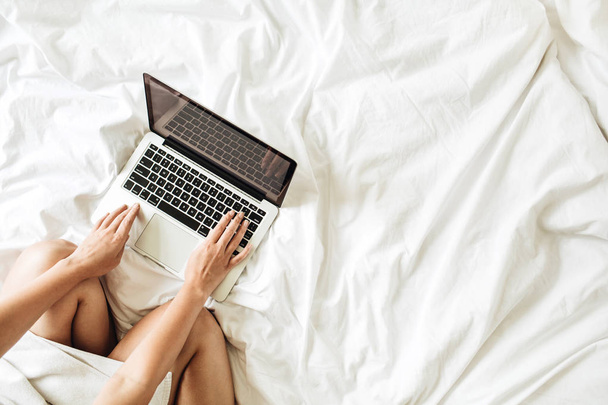 Working at home concept. Girl is working on her laptop in bed with white linens. Flat lay, top view minimalistic workspace background. Relaxing working process.  - Photo, Image