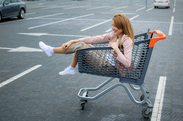 Ciemny szary i pomarańczowy koszyk z piękną dziewczyną wewnątrz stoi na parkingu w supermarkecie. - Zdjęcie, obraz