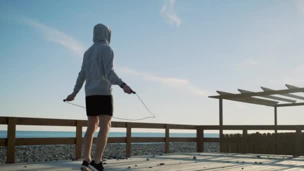 Athletic man is working out on seashore in daytime, jumping rope alone - Footage, Video
