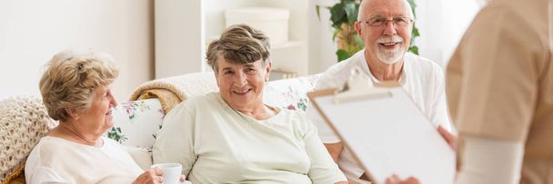 Vista panorâmica de idosos felizes sentados em um sofá no centro de cuidados e olhando para sua enfermeira
 - Foto, Imagem