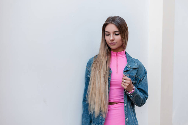ziemlich glamouröse junge blonde Frau in einem modischen rosafarbenen Sommer-Sportanzug in einer stylischen blauen Jeansjacke, die eine Pause in der Stadt genießt. schönes nettes Mädchen Mode-Modell steht in der Nähe der weißen Wand. - Foto, Bild