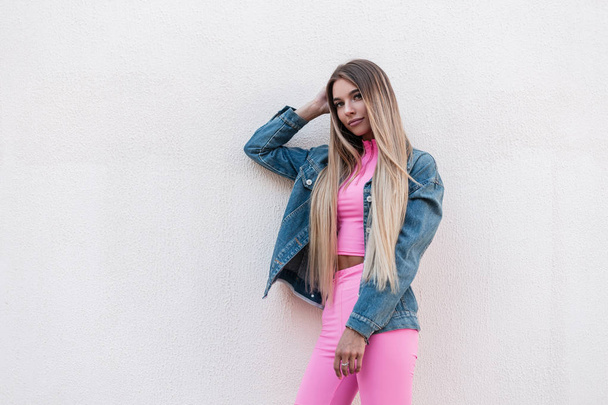 Modern urban young woman with chic long hair in a blue fashionable denim jacket in a glamorous pink sports suit poses near a building in the city. European stylish blonde girl outdoors. Retro style. - Photo, Image