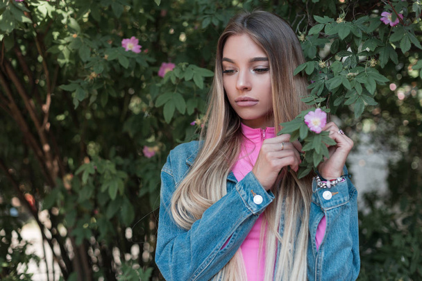 Pretty beautiful young woman with long hair with natural make-up in a vintage pink top in a stylish denim jacket posing standing near a flowering bush with pink flowers. Cute girl resting in the park. - Fotografie, Obrázek