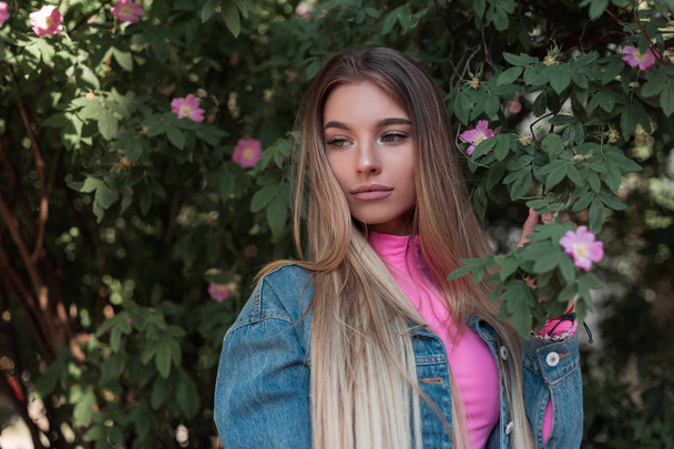 Jolie jolie jeune femme aux cheveux longs luxueux dans une veste en denim vintage dans un élégant haut rose près d'un buisson fleuri vert dans le jardin. Belle fille glamour se détend à l'extérieur un jour d'été
 - Photo, image