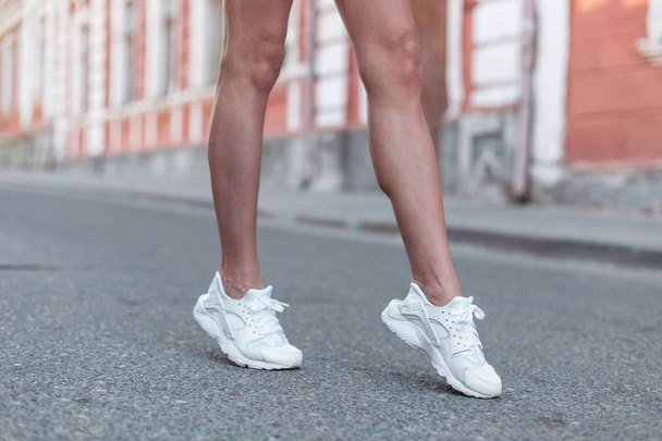 Moderne jonge vrouw met slanke mooie benen in modieuze witte sneakers loopt langs de straat. Stijlvolle sportieve Damesschoenen. Zomer stijl. Close-up van vrouwelijke benen met schoenen. - Foto, afbeelding
