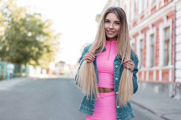 Θετική νεαρή γυναίκα με ένα όμορφο χαριτωμένο χαμόγελο σε ένα μοντέρνο ντένιμ μπουφάν σε ένα κομψό ροζ πουκάμισο με σορτς στέκεται στο δρόμο στην πόλη και κρατώντας τα μαλλιά. Ελκυστική ξανθιά κορίτσι που ποζάρει. - Φωτογραφία, εικόνα