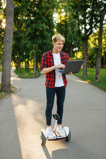 Jeune homme chevauchant sur mini gyroscope avec ordinateur portable, parc d'été. Loisirs de plein air avec gyroboard électrique. Eco transport avec technologie d'équilibrage, véhicule gyroscope électrique
 - Photo, image