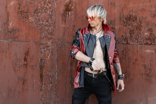 Sexy young man with blond hair in trendy red glasses in a jacket with jeans with a vintage gun poses near an old rusty wall. Handsome guy hipster. Old fashioned style. - Valokuva, kuva