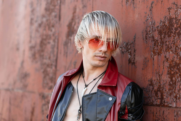 Portrait of a young modern hipster man in a trendy black-red leather jacket in vintage red glasses with a stylish hairstyle outdoors near a rusty metal wall.   - Fotó, kép