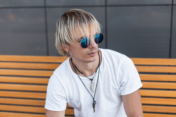 Jeune homme branché en lunettes de soleil bleues en t-shirt blanc assis sur un banc en bois près d'un bâtiment gris un jour d'été. Modèle de gars attrayant à l'extérieur. Collection tendance été hommes
. - Photo, image