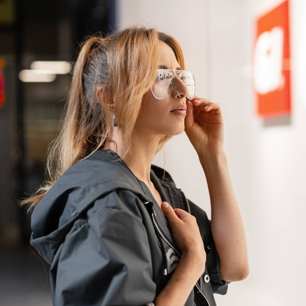 Young sexy hipster woman with red hair in a vintage hooded jacket in stylish glasses stands near a bright glass showcase in a mall. American attractive girl travels around the store. Street style. - Φωτογραφία, εικόνα