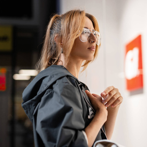 American young woman with a trendy hairstyle in a stylish jacket with hood in glasses standing near modern bright glass wall in shop. Redhead pretty urban girl model. Fashionable women's clothing. - Фото, изображение