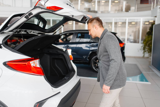 Man looks at the trunk of new car in showroom. Male customer choosing vehicle in dealership, automobile sale - Photo, Image